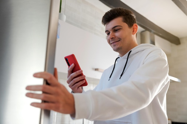 Caucasian man smiling in the kitchen making the shopping list He is checking the shopping list on his mobile phoneHe is dressed in a white sweatshirt and his mobile phone is redEuropean Spain