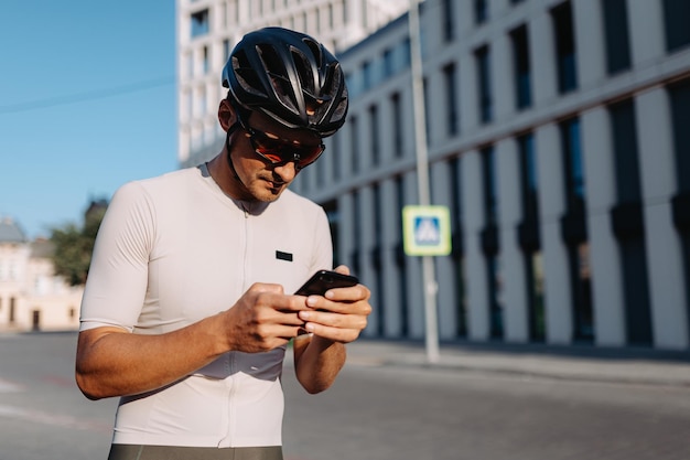 Caucasian man in sport clothes helmet and glasses texting on cell phone while standing on city street Cyclist resting outdoors after morning workout