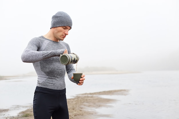Caucasian man standing on bank of river in foggy day and drinking tea
