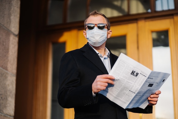 Caucasian man in sunglasses, medical mask and formal black suit, stands near building outdoor, holds newspaper in hands