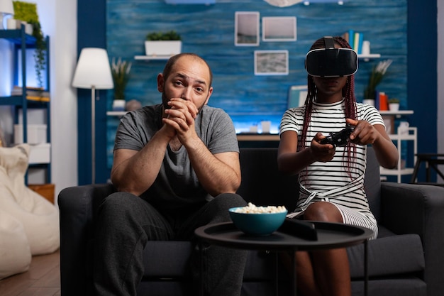 Caucasian man watching african american woman playing VR game. Husband sitting besides wife using virtual reality goggles. Diverse couple enjoying wearable technology and simulation games