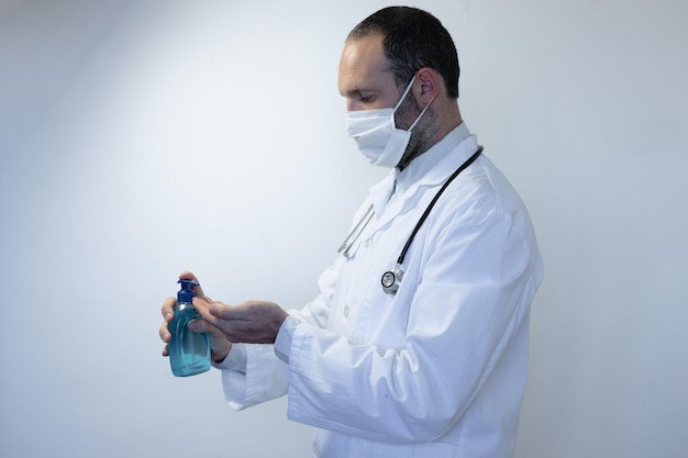 Caucasian man, wearing doctor uniform and a stethoscope on his shoulders, wearing a face mask and standing in an empty white room, disinfecting his hands with sanitizer