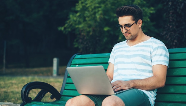 Uomo caucasico che indossa occhiali da vista è seduto su una panchina in un parco e utilizza un computer portatile