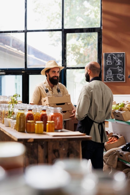 Caucasian man wearing a hat and holding boxes is interacting with male shopkeeper in grocery store filled with eco friendly and organic food products Farmer carrying packages while talking to vendor