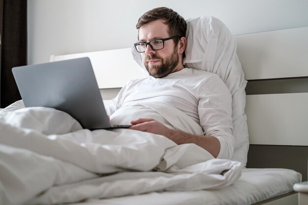 Caucasian man in white pajamas works with the laptop remotely while sitting in bed in the morning