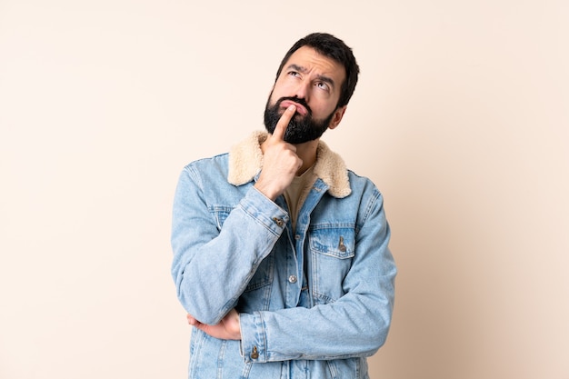 Caucasian man with beard over isolated wall having doubts while looking up