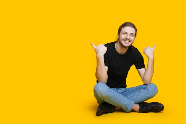 Caucasian man with beard and long hair sitting on the floor and gesturing the like sign near the yellow studio free space