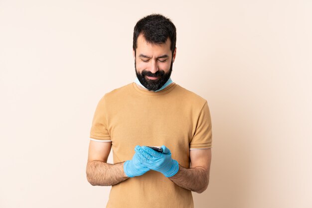 Uomo caucasico con la barba che protegge dal coronavirus con una maschera e guanti sul muro inviando un messaggio con il cellulare