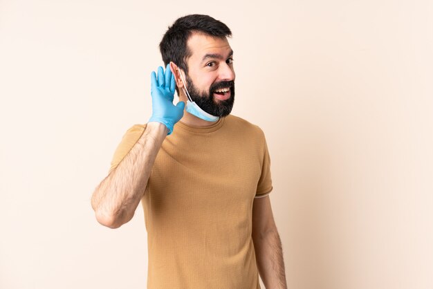 耳に手を置くことによって何かを聞いて孤立した壁にマスクと手袋で保護するひげを持つ白人男