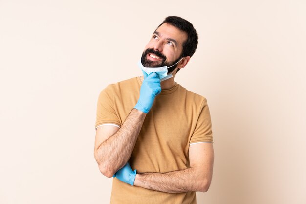 Caucasian man with beard protecting  with a mask and gloves over isolated wall and looking up