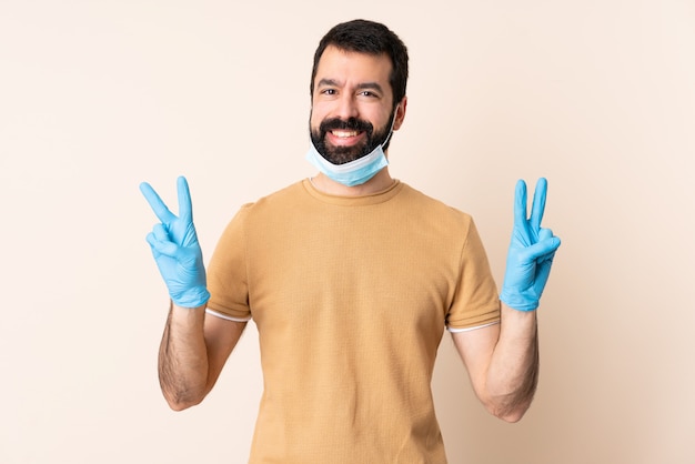 Caucasian man with beard protecting  with a mask and gloves over isolated wall showing victory sign with both hands