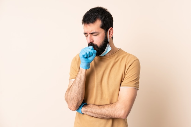 Uomo caucasico con la barba che protegge con una maschera e guanti sopra la parete che tossisce molto