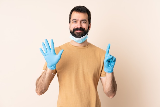 Кавказский мужчина с бородой, защищая маску и перчатки на стену, считая шесть с пальцами