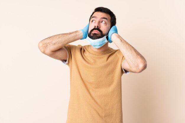 Caucasian man with beard protecting with a mask and gloves over wall frustrated and covering ears