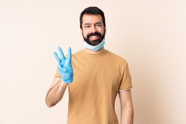 Caucasian man with beard protecting with a mask and gloves over wall happy and counting three with fingers