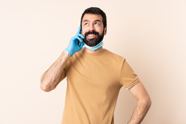 Caucasian man with beard protecting with a mask and gloves over wall keeping a conversation with the mobile phone