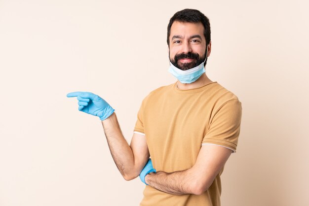 Caucasian man with beard protecting with a mask and gloves over wall pointing finger to the side