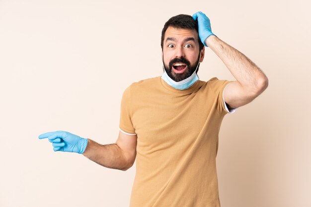 Caucasian man with beard protecting with a mask and gloves over wall surprised and pointing finger to the side