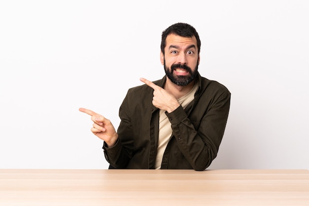 Caucasian man with beard in a table frightened and pointing to the side.