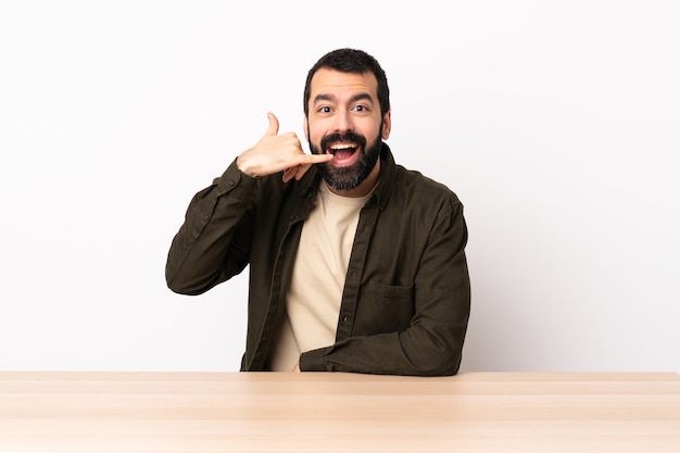 Caucasian man with beard in a table making phone gesture. Call me back sign.