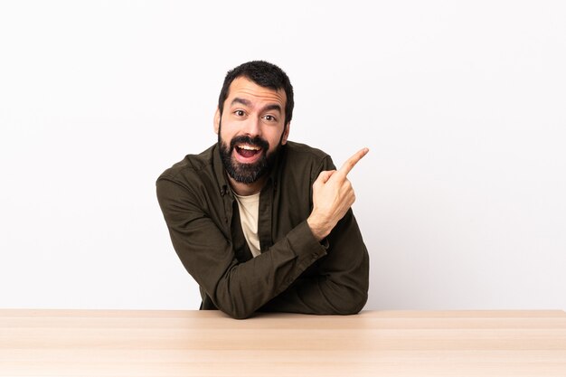 Caucasian man with beard in a table surprised and pointing side.