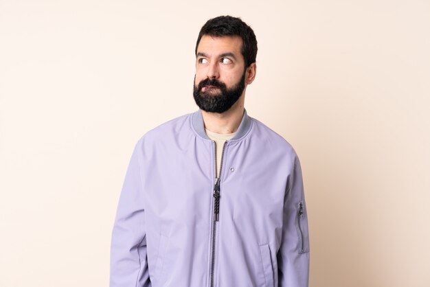 Caucasian man with beard wearing a jacket over isolated space having doubts while looking up