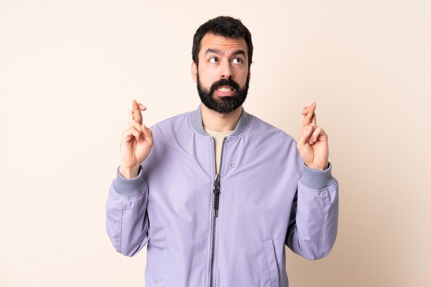 Caucasian man with beard wearing a jacket over isolated wall with fingers crossing and wishing the best