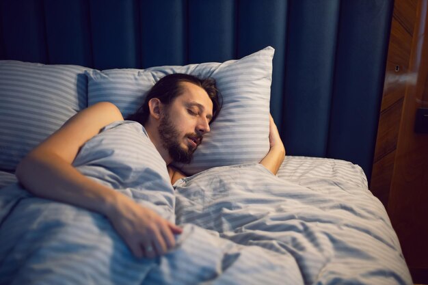 Un uomo caucasico con la barba in una maglietta bianca è sdraiato sotto una coperta e dorme in un letto