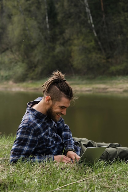 Caucasian man with dreadlocks and beard lying in clearing on river bank in spring park and working on laptop Happy and smiling Concept of remote work and business using Internet technologies