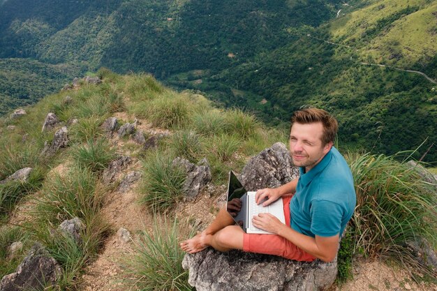 Caucasian man with laptop sitting on the edge of a mountain with stunning views working on laptop Freelancer in Sri Lnaka