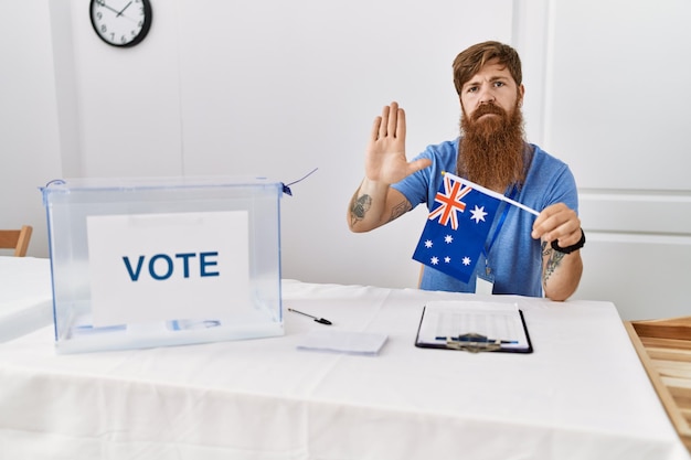 Caucasian man with long beard at political campaign election holding australia flag with open hand doing stop sign with serious and confident expression, defense gesture