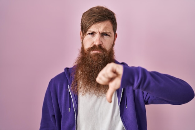 写真 ピンクの背景の上に立っている長いあごひげを生やした白人男性は、不幸で怒っているように見え、拒絶と親指を下に向けたネガティブなジェスチャーを示しています。悪い表現。