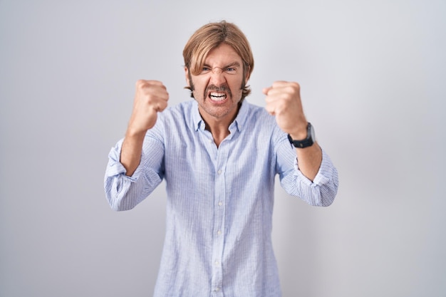 Caucasian man with mustache standing over white background angry and mad raising fists frustrated and furious while shouting with anger. rage and aggressive concept.