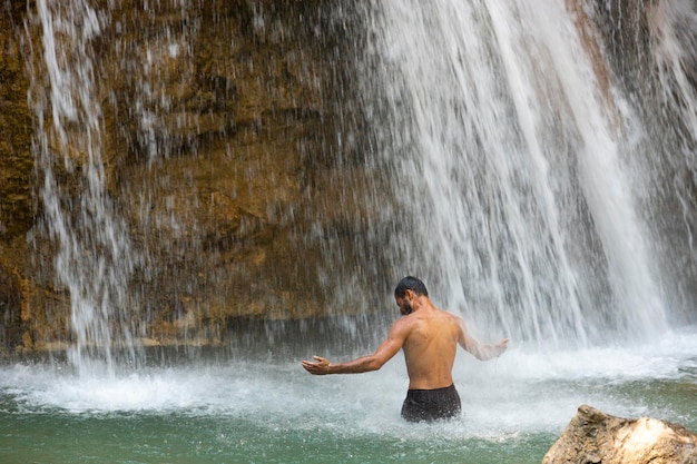 Caucasian man with raised arms and standing with his back enjoying and bathing in a fresh mountain waterfall on a mountain river in Europe. High quality photo