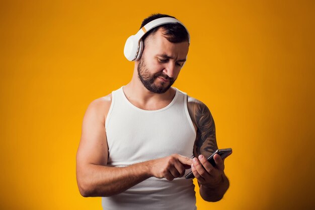 Фото Белый мужчина с татуировкой на плече слушает музыку в наушниках выбирает звуковую дорожку плейлист песни подкаст на телефоне на желтом фоне люди и концепция образа жизни