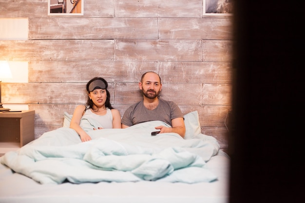 Caucasian married couple in pajamas watching tv laying on cozy bed.