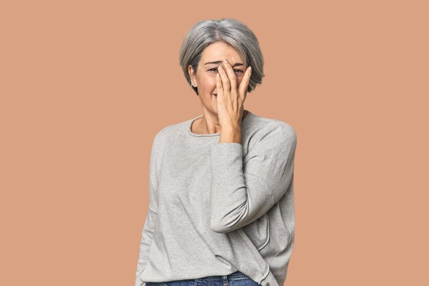 Photo caucasian midage female on studio background blink at the camera through fingers