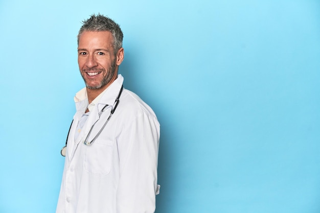 Caucasian middleaged doctor on blue background looks aside smiling cheerful and pleasant