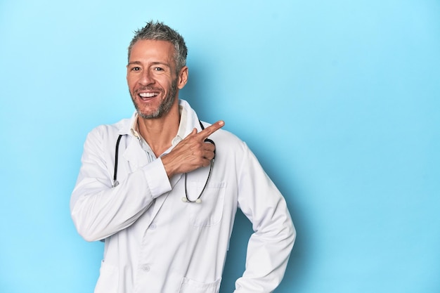 Caucasian middleaged doctor on blue background smiling and pointing aside showing something at blank space