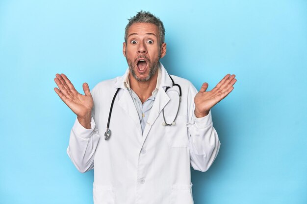 Caucasian middleaged doctor on blue background surprised and shocked