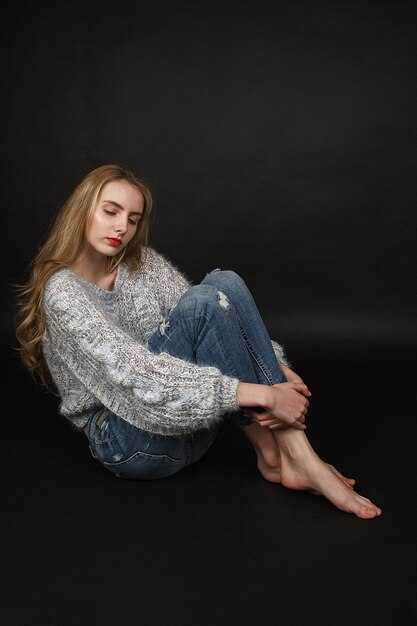 caucasian natural young pretty girl with long blonde hair in  sweater and jeans sitting on the floor