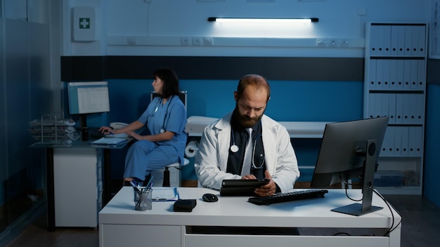病院のオフィスでヘルスケア治療の計画を開始する前に、患者の病気の専門知識を検索するタブレット コンピューターを保持している白人の医師。夜遅くまで働く医療チーム