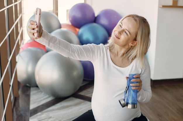 Caucasian pregnant woman in third trimester make a selfie and holding a bottle with water Woman in bright fitness studio with big windows Blonde woman wearing whit shirt and black leggins