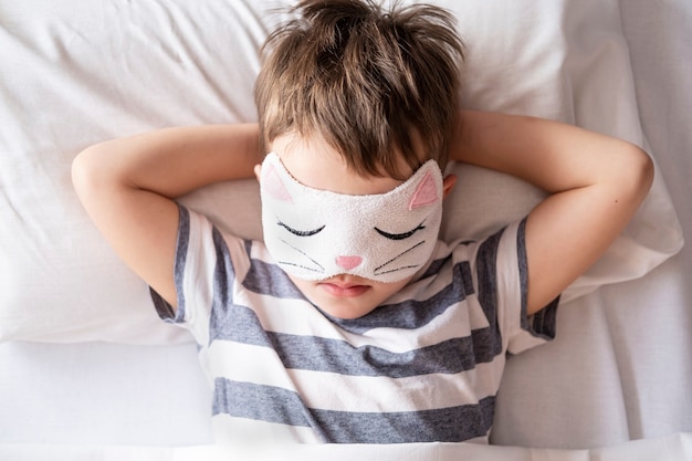 Caucasian preschool boy in kitty eye mask striped pyjamas lying in white bed.