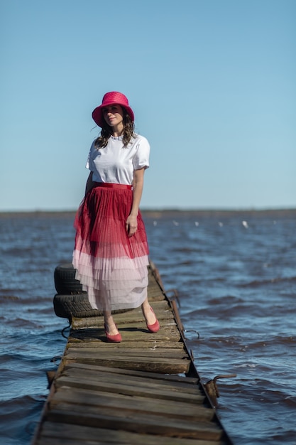 Foto donna caucasica piuttosto di mezza età in posa su un molo con una gonna rosa e un cappello a secchiello in una giornata di sole in russia