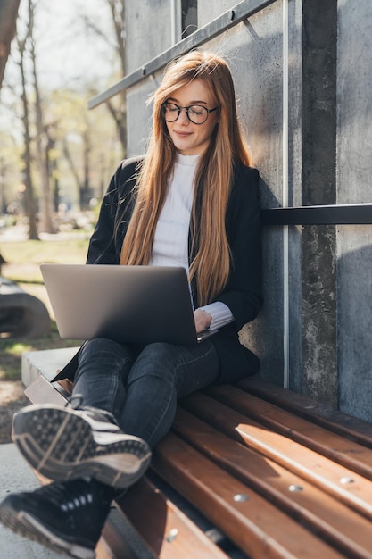 Caucasian professional businesswoman sitting on a bench with computer laptop working outside in the city park