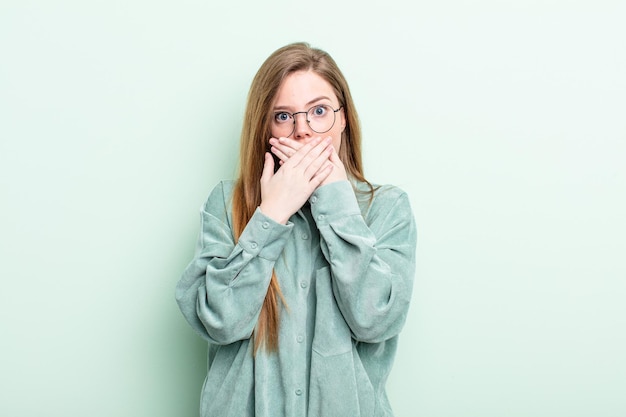 Donna caucasica dai capelli rossi che copre la bocca con le mani con un'espressione scioccata e sorpresa, mantenendo un segreto o dicendo oops