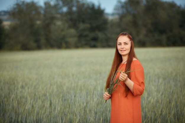 Donna dai capelli rossi caucasica in un vestito rosso che cammina su un campo di fattoria con grano al tramonto su un raccolto di cibo biologico estivo dayfuturo