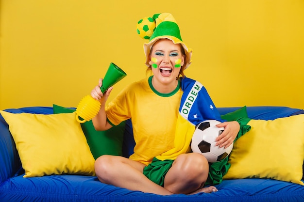 Foto tifoso di calcio del brasile della donna della testarossa caucasica sul divano con il tifo della palla e del corno