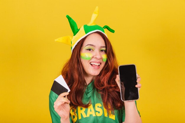 Кавказская рыжеволосая женщина, бразильская футбольная фанатка, держащая кредитные карты и концепцию мобильных продаж мобильного телефона